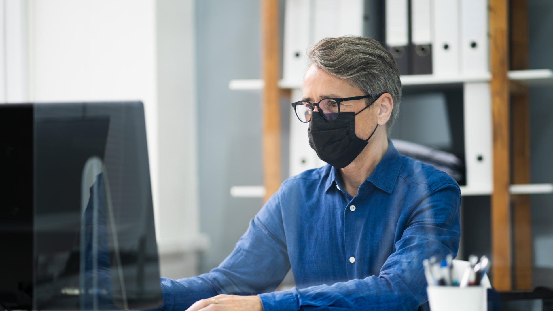 Toimistossa työskentelevä mies on suojatunut maskilla.