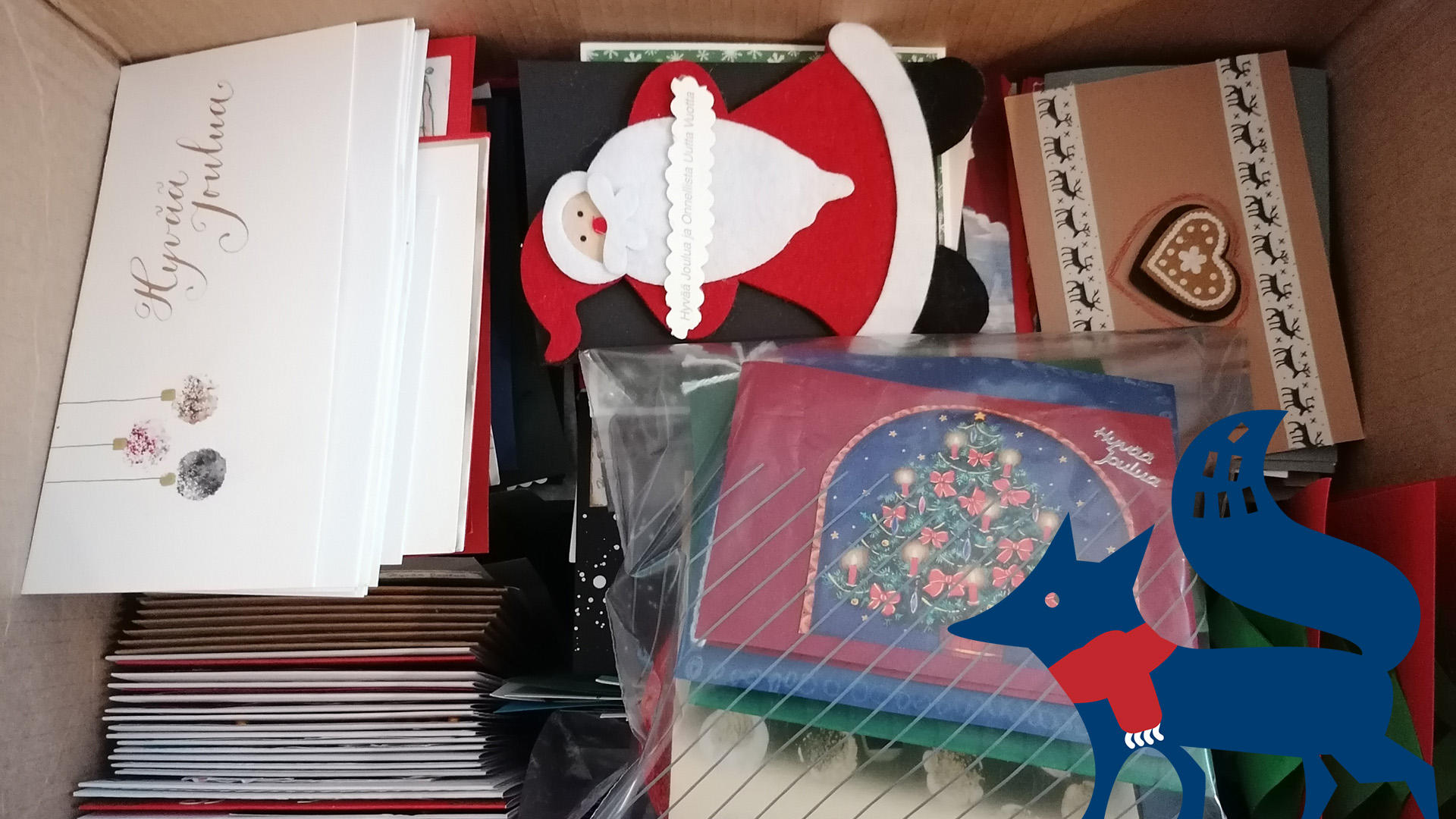 Paljon joulukortteja laatikossa. Alakulmassa sininen kettu, jolla on jouluinen, punainen kaulaliina.
