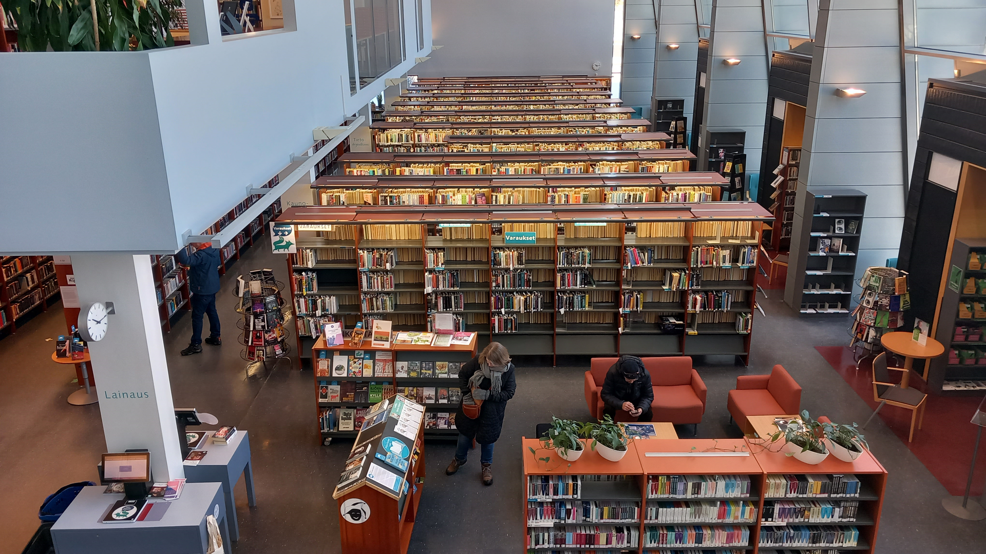 Kirjastosalin näkymässä kirjahyllyjä sekä kolme asiakasta.