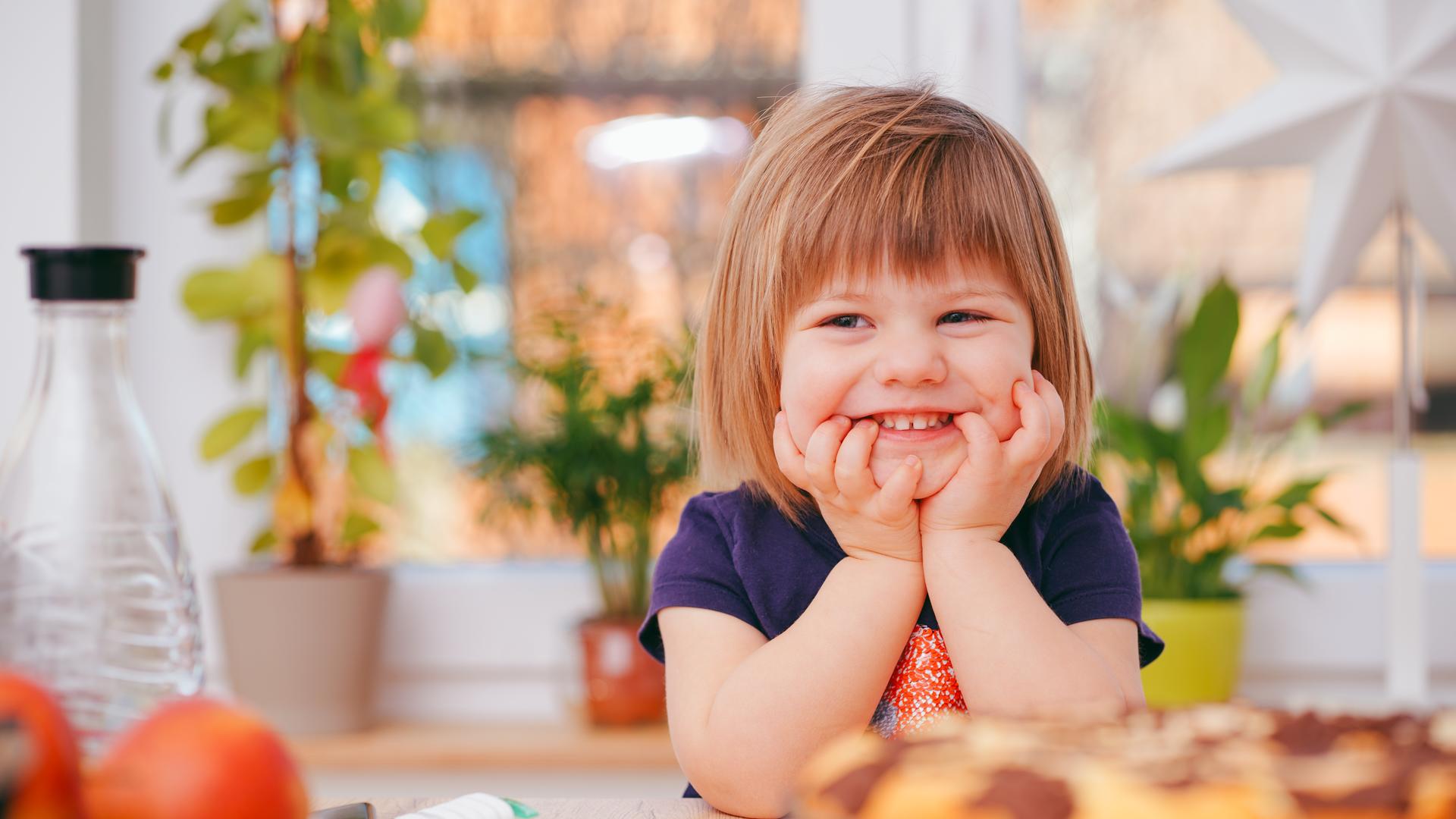 Pikkutyttö nojaa käsiinsä ja hymyilee ruokapöydän ääressä.