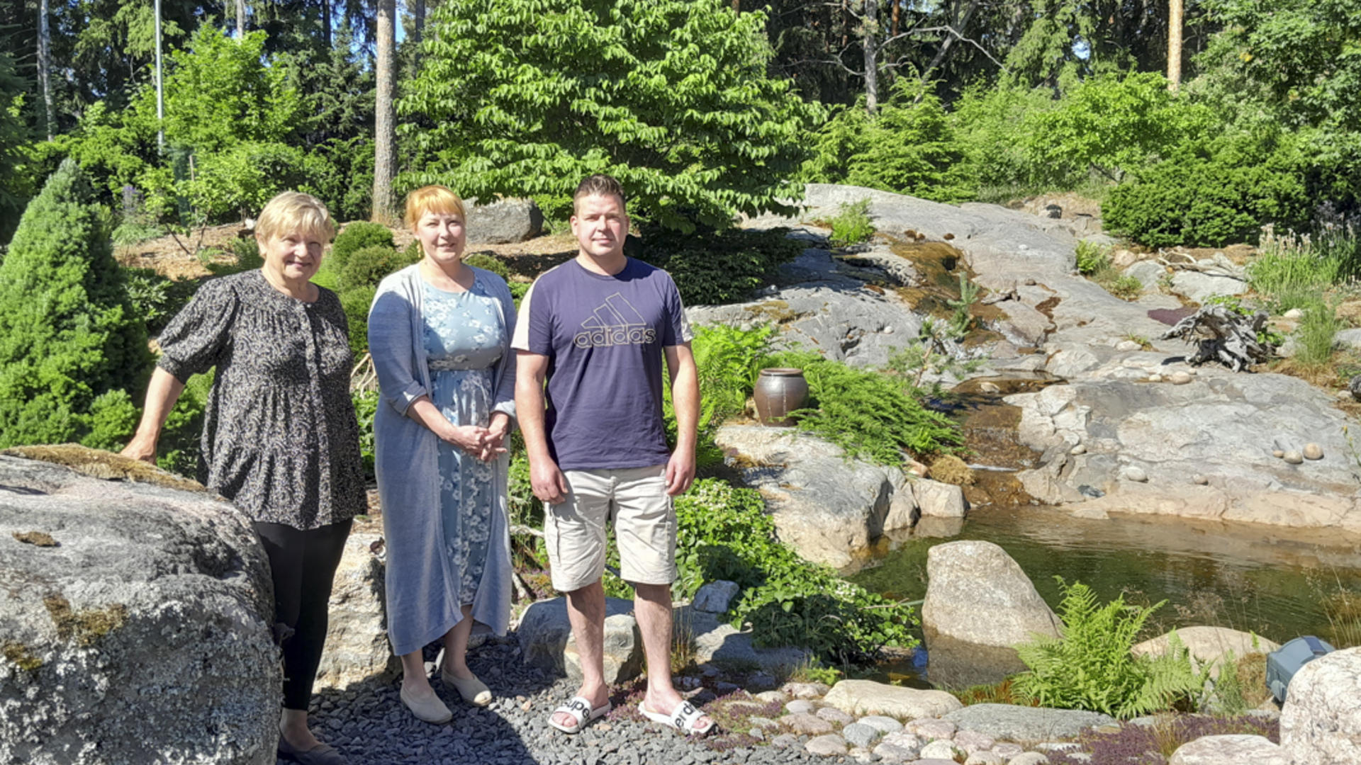 Anne Hanhiniemi, Mari Salola ja Jari Silventoinen seisovat puutarhassa vieressään iso kivi ja vesiallas.
