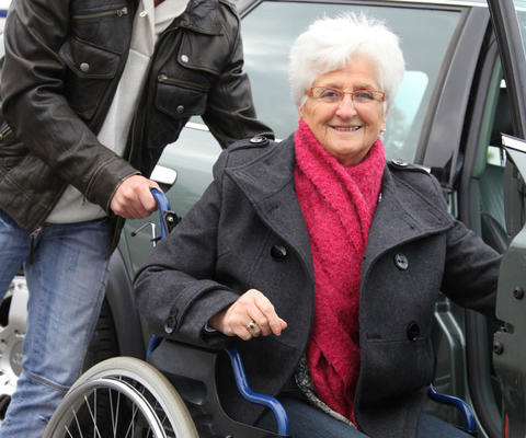 Avustaja auttaa pyörätuolissa istuvaa naista autoon.