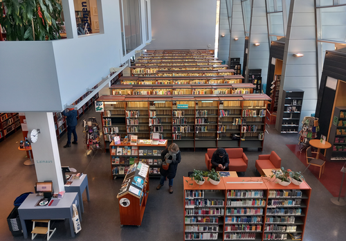 Kirjastosalin näkymässä kirjahyllyjä sekä kolme asiakasta.