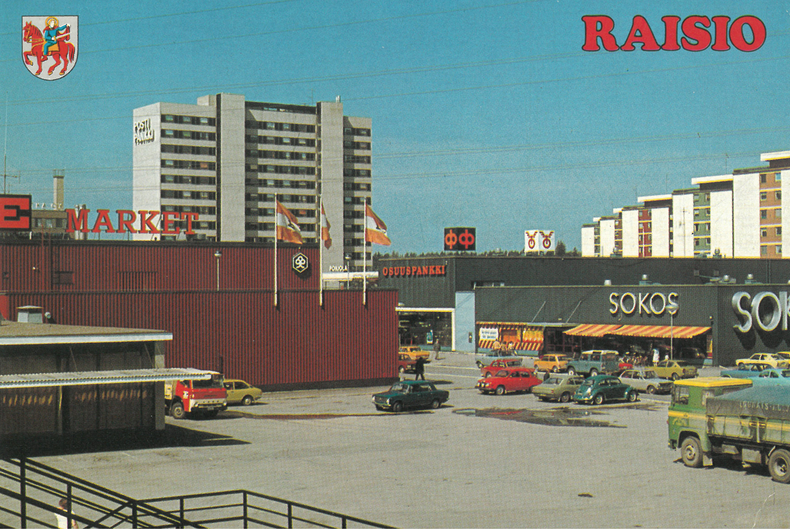 Postikortti 1970-luvulta. Kuvassa Raision keskustan kaupparakennuksia ja parkkipaikkaa.