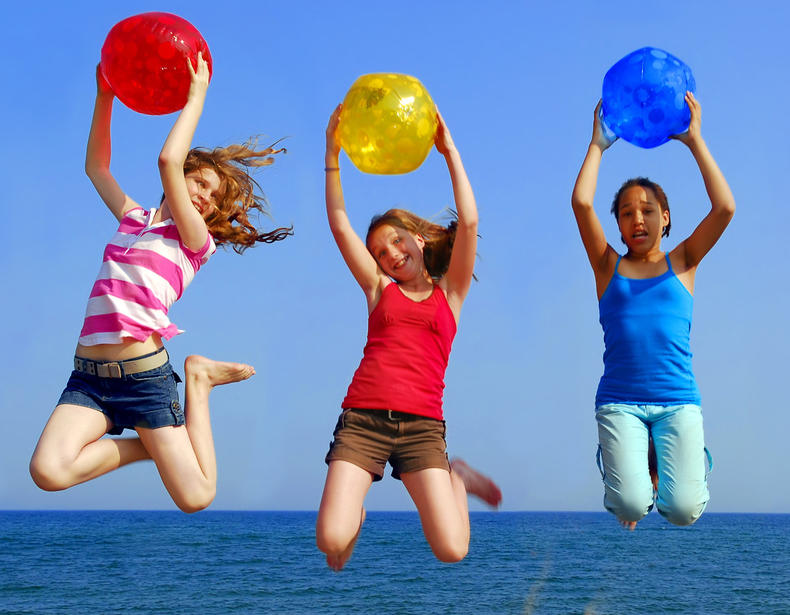 Kolme tyttöä hyppää rannalla ilmaan pitäen värikkäitä rantapalloja käsissään.